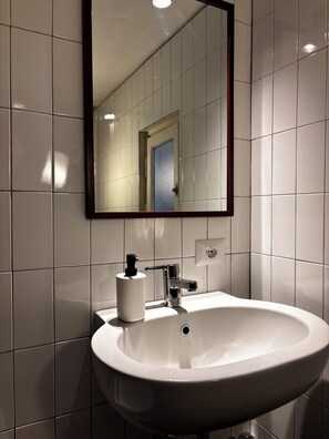 Baño con azulejos blancos, un lavamanos blanco, un espejo y una puerta.