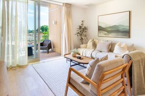 Ein modernes Wohnzimmer mit einer Couch, einem Couchtisch und einem Holzsessel. Eine Schiebetür aus Glas führt in einen Außenbereich mit einem Sessel und einem Tisch. An der Wand hängt ein Gemälde, und auf einem Beistelltisch steht eine Pflanze.