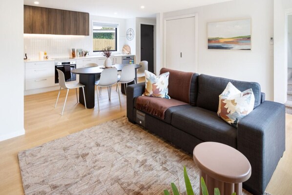 Uma sala de estar moderna com um sofá cinza escuro, uma mesa de centro redonda e uma área de jantar com uma mesa e cadeiras brancas.