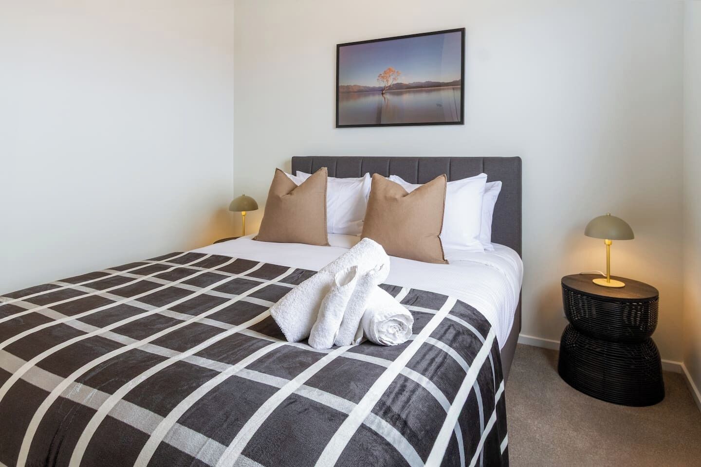 A neatly made bed with a checkered bedspread, two pillows, and a rolled towel. There is a framed picture on the wall above the bed.