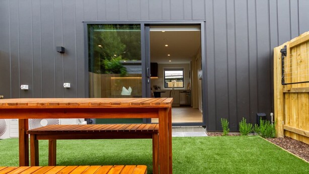 Une maison moderne avec une table et un banc en bois au premier plan, une porte-fenêtre coulissante donnant sur une cuisine et un petit jardin.