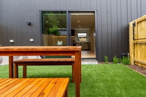 Uma casa moderna com uma mesa e banco de madeira em primeiro plano, uma porta de correr de vidro que dá para uma cozinha, e uma pequena área de jardim.