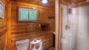 Baño de cabaña de madera con inodoro, un estante pequeño, una ventana y ducha.