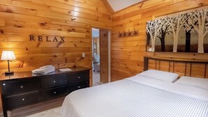 A wooden-paneled room with a bed, a dresser, a lamp, and a wall-mounted artwork. - Springwood Cottage Resort - Cottage 5 (Arden)