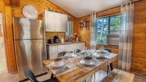 Cozinha de cabana de madeira com geladeira de aço inoxidável, armários brancos e uma mesa de jantar posta com pratos, copos e talheres.