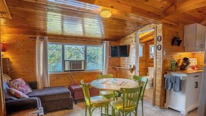 A cozy cabin interior with a wooden ceiling, a dining area with a table and chairs, a sofa with cushions, a small kitchen with a refrigerator, and a TV mounted on the wall. - Springwood Cottage Resort - Cottage 8 (Arden)