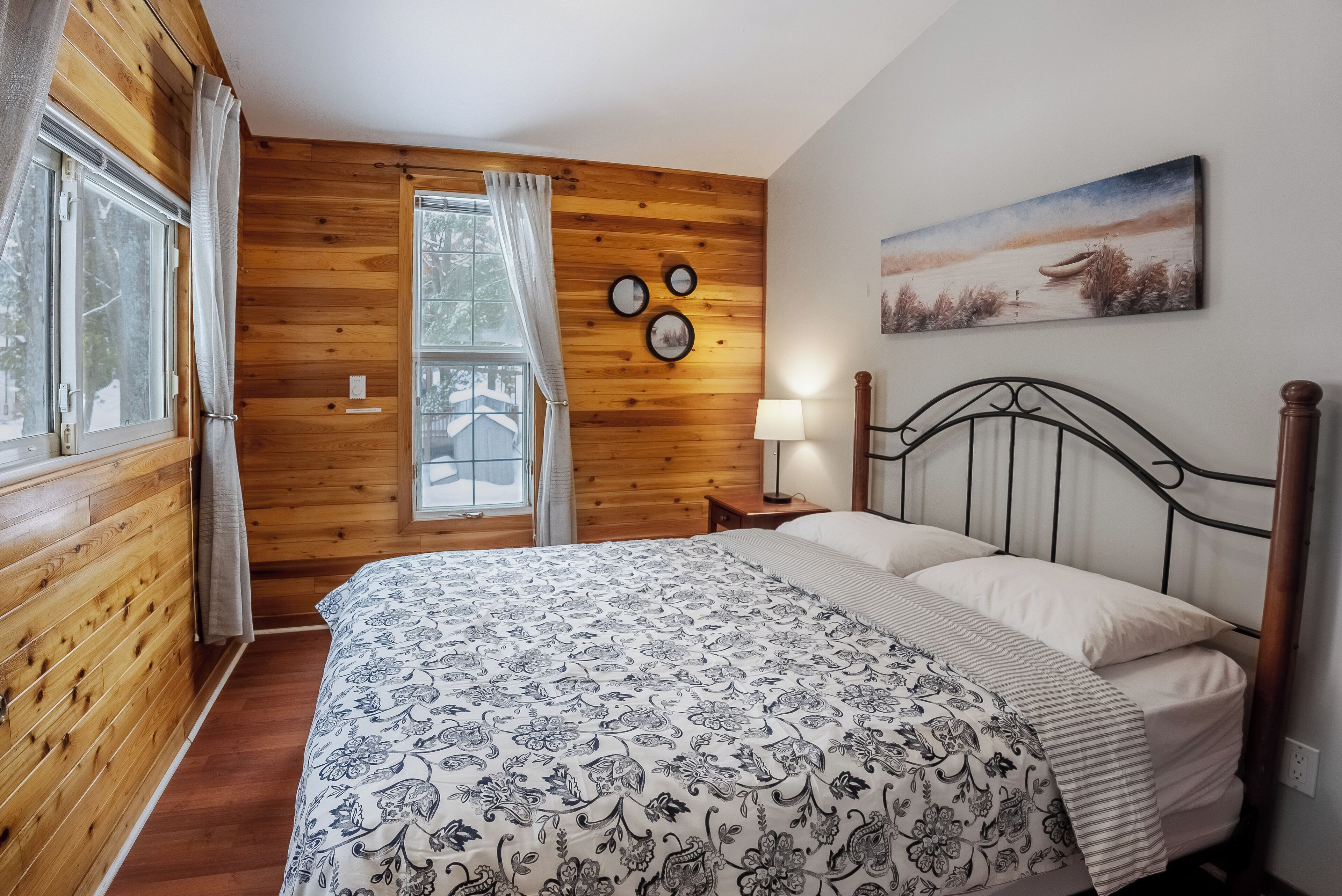 Een slaapkamer met houten lambrisering, een bed, nachtkastje, lamp en schilderij aan de muur.