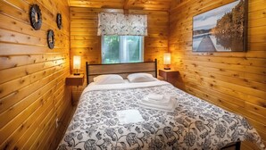 Un dormitorio de cabaña de madera con una cama, dos lámparas de noche, una ventana con persianas y un cuadro en la pared.