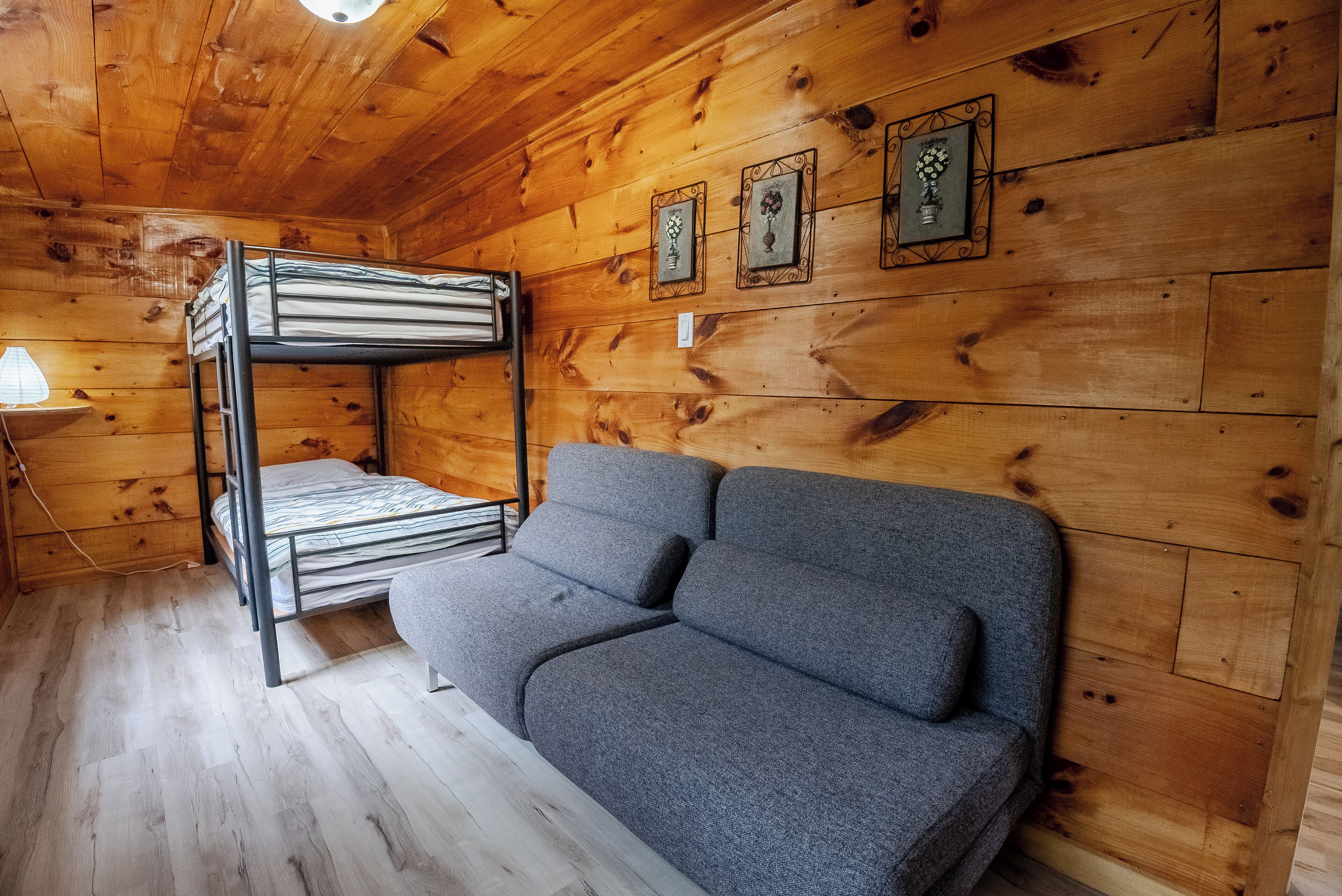 A wooden cabin room with a bunk bed, a gray sofa, and three wall-mounted picture frames.