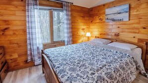 A wooden cabin bedroom with a bed, a window with curtains, a lamp, and a painting on the wall. - Springwood Cottage Resort - Cottage 15 (Arden)