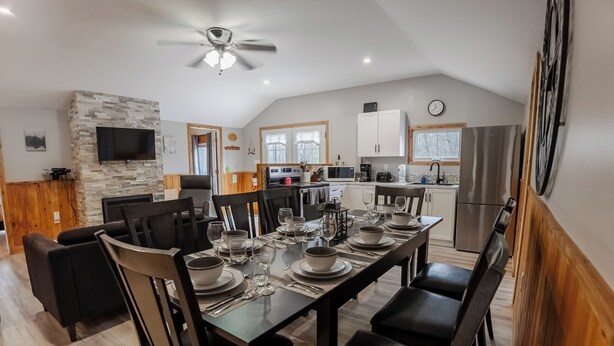A modern kitchen with a stone accent wall, a dining area with a large table set for six, and a ceiling fan. - Springwood Cottage Resort - Cottage 15 (Arden)