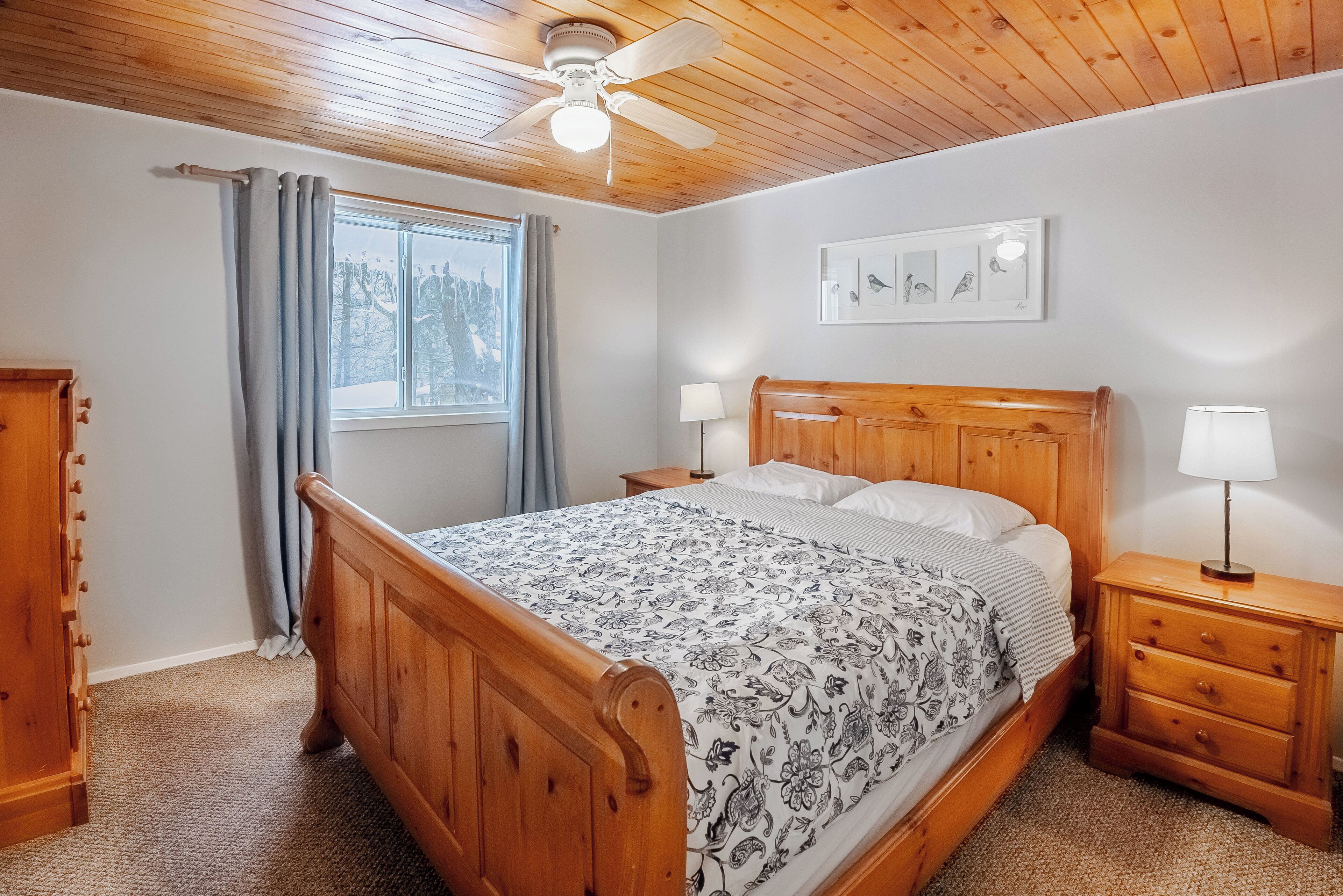 Un dormitorio con cama de madera, una mesita de noche, un aparador, un ventilador de techo y una ventana con cortinas.