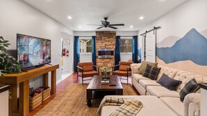 Un salon moderne avec un téléviseur posé sur un meuble en bois, une cheminée en pierre, deux fauteuils en cuir, un canapé beige et un ventilateur de plafond.