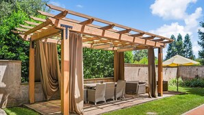 A wooden pergola with a table and chairs, surrounded by greenery. - The Hideaway | Pool,Tennis Court, Outdoor Kitchen (Santa Rosa)
