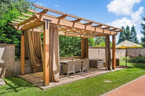 Eine hölzerne Pergola mit Tisch und Stühlen, umgeben von Grünpflanzen.