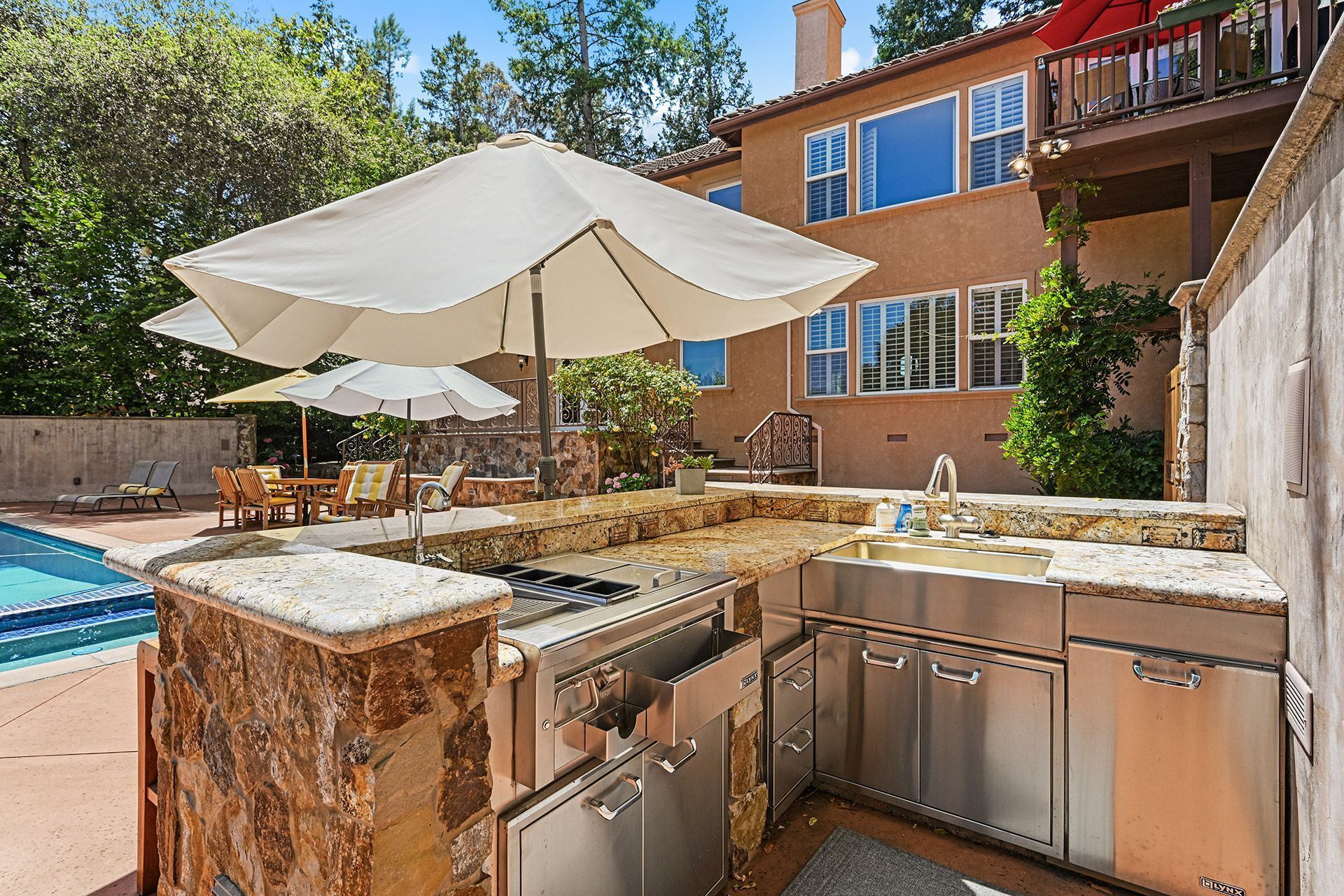 An outdoor kitchen with a stone countertop, stainless steel appliances, and a pool in the background.
