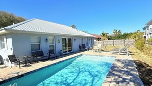 Una piscina rectangular con un borde de baldosas, una casa blanca con techo de metal y una cerca de madera al fondo.