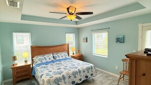 Un dormitorio con cama, ventilador de techo, mesita de noche, cómoda y ventana con persianas.
