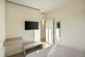 Une chambre moderne avec un lit, un téléviseur fixé au mur, une porte donnant sur une autre pièce et un petit banc.