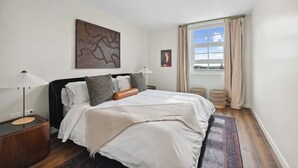 Una camera da letto con un letto, un comodino, una lampada, una finestra con tende e un quadro appeso al muro.