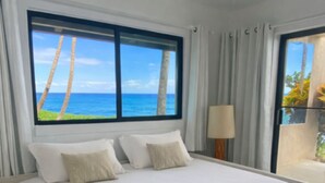 Ein großes Bett mit weißen Leinen und vielen Kissen, ein Deckenventilator und Meerblick durch ein großes Fenster.