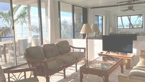 Ein großzügiges Wohnzimmer mit einer großen Couch, einem Couchtisch und einem Fernseher. Deckenventilatoren und große Fenster bieten Blick auf den Ozean.