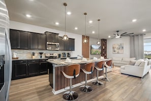 A modern kitchen with dark cabinets, a central island with bar stools, and a dining area with a white sofa. - 10br Pool Retreat | Theatre • Arcade | Sleeps 24 (davenport)