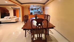 Un espace repas avec une longue table en bois et des chaises, un canapé avec des coussins, et une cuisine en arrière-plan.