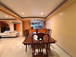 A dining area with a long wooden table and chairs, a sofa with cushions, and a kitchen in the background.