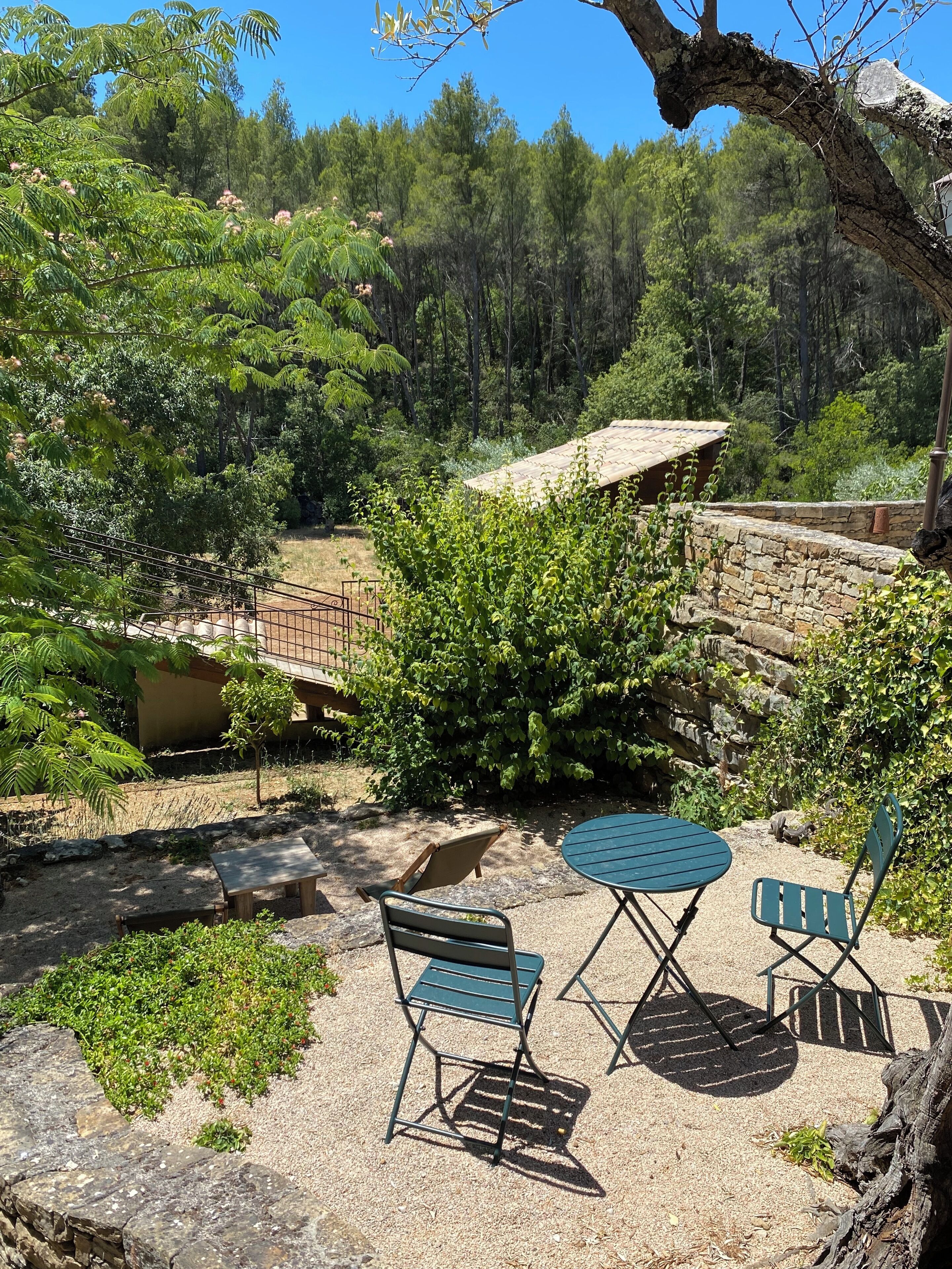 Un espace extérieur aménagé avec deux tables et quatre chaises, entouré de verdure et d’un mur en pierre.
