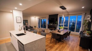 A modern kitchen with a central island, dining area with chairs, a fireplace, and large windows with curtains. - 206 | Bright Modern Condo | Pool and River View (Baie-Saint-Paul)