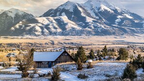 A rustic cabin with a snow-covered roof, surrounded by a mountainous landscape and a few scattered houses. - Adorable Montana Cabin by Yellowstone! (Emigrant)