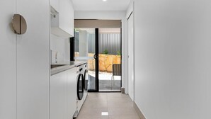 A modern kitchen with a washing machine, a countertop, and a sliding glass door leading to an outdoor area with a stone wall and a chair. - Marys Tide & View Vista at Long Beach (Long Beach)