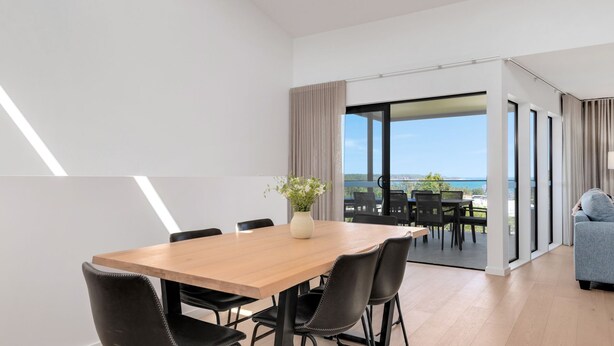 A modern dining area with a wooden table and black chairs, a vase with flowers, and a view of the sea through large glass doors. - Marys Tide & View Vista at Long Beach (Long Beach)