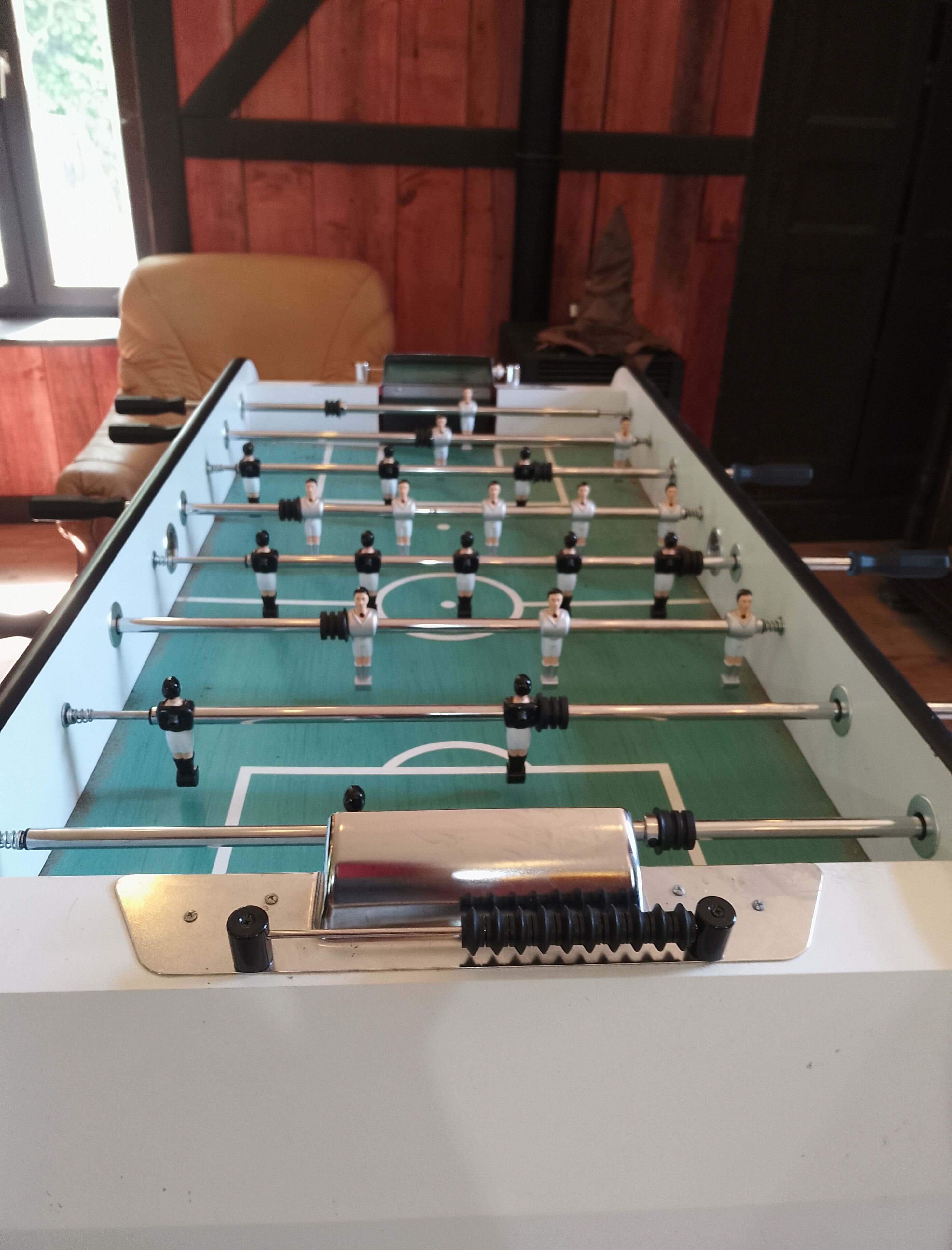 Una mesa de fútbol de mesa con superficie de juego verde y barras de metal, frente a una pared de madera.