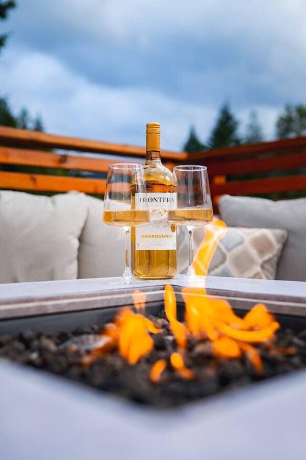 A bottle of wine and two glasses on a table with a fire pit in the foreground.