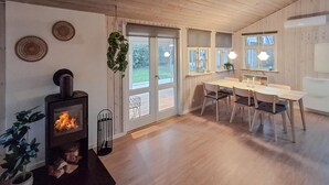 A cozy living room with a wood-burning stove, a dining area with a table and chairs, and a large window offering a view of the outdoors. - Lovely home in Sjølund with WiFi (Sjølund)