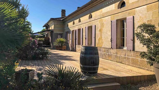 Un patio con una cubierta de madera, un barril grande y un edificio con ventanas de arco y contraventanas.