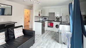 Un apartamento moderno con sala de estar que cuenta con un sofá de cuero negro y una mesa de comedor. La cocina incluye armarios blancos, un microondas y una lavadora.