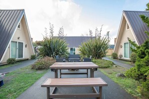 Une cour avec une table en bois et des bancs, entourée de deux bâtiments aux toits inclinés et aux grandes fenêtres.