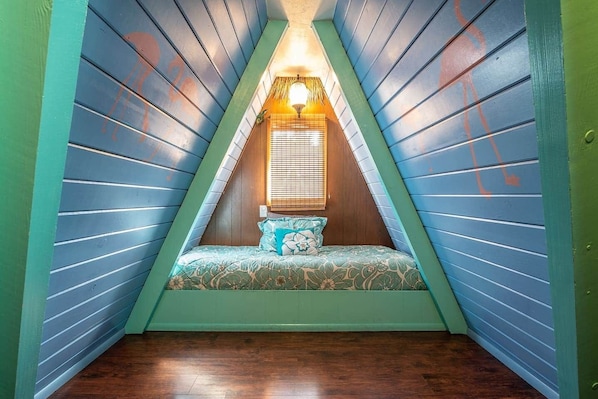 Un dormitorio ático pequeño y triangular, con techo inclinado, una cama individual con ropa de cama floral y una ventana con persianas.