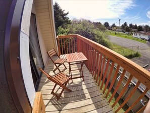 Une terrasse en bois avec une table et deux chaises, une rambarde en métal, et une vue sur un parking et des bâtiments en arrière-plan.