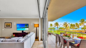 A balcony with a view of palm trees and the ocean, a sofa, a TV, and a dining table set for two. - Honua Kai | Ocean View 8 BR, Sleeps 22 | Car Incl. w/ 6+ Nights | HKH ML-2594 by KBM (Lahaina)