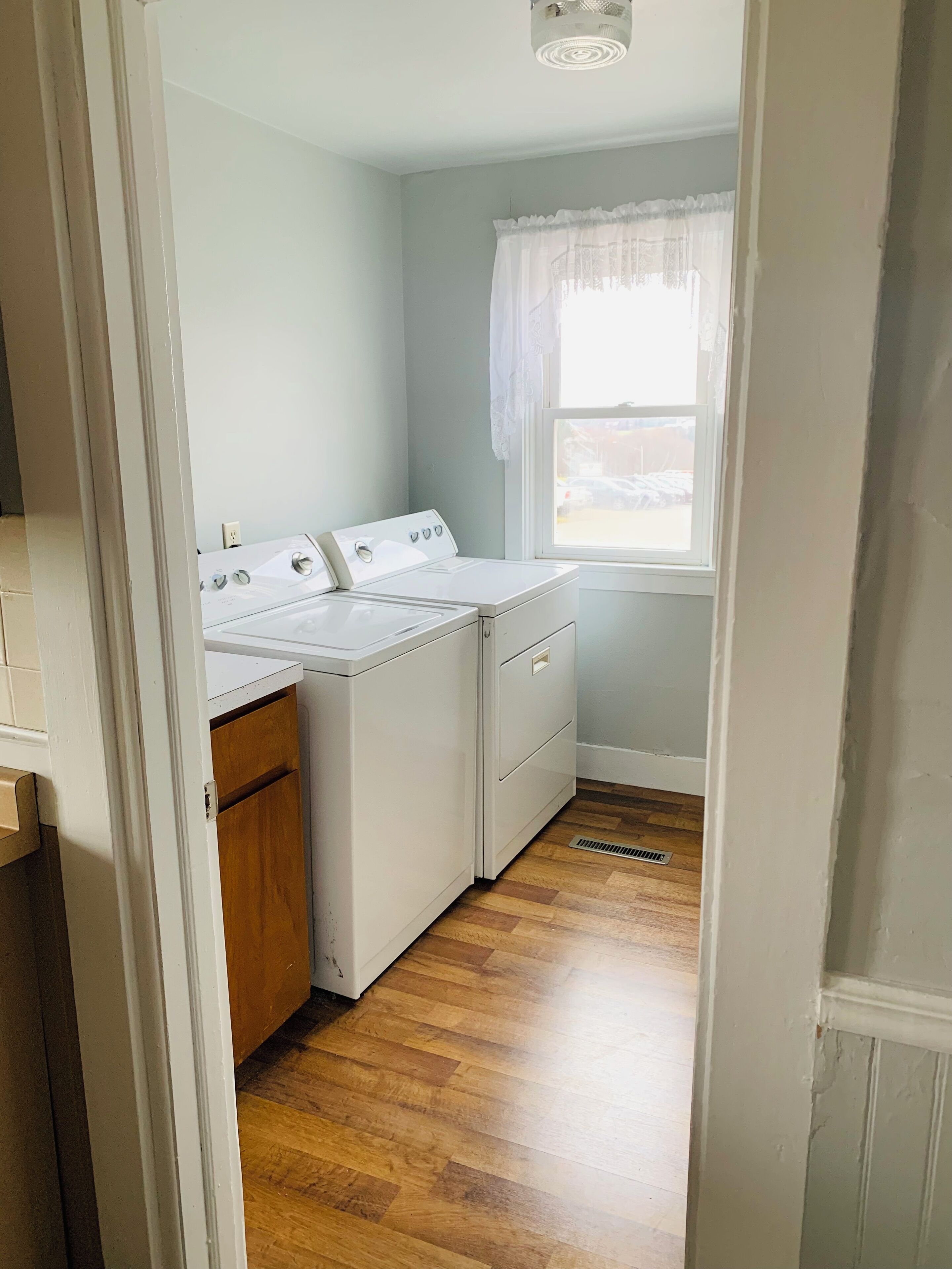 Une buanderie équipée d’un lave-linge et d’un sèche-linge, avec un plancher en bois et une fenêtre agrémentée d’un rideau blanc.