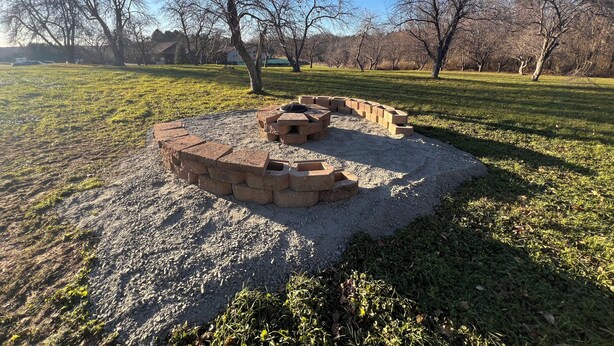 Un pozo de fuego circular de piedra con una mesa metálica en el centro, rodeado por un muro bajo de piedra en un parque con césped y árboles al fondo.