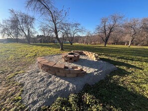 A circular stone fire pit with a metal table in the center, surrounded by a low stone wall in a grassy park area with trees in the background. - Adorable 3-bedroom house with scenic views in quaint Neillsville, WI (Neillsville)