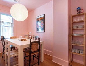 A dining area with a white table, wooden chairs, a hanging light fixture, and a wall-mounted shelf with dishes.