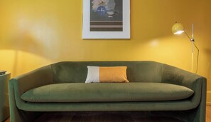 A green sofa with a white and orange pillow, a framed picture on the wall, and a floor lamp.