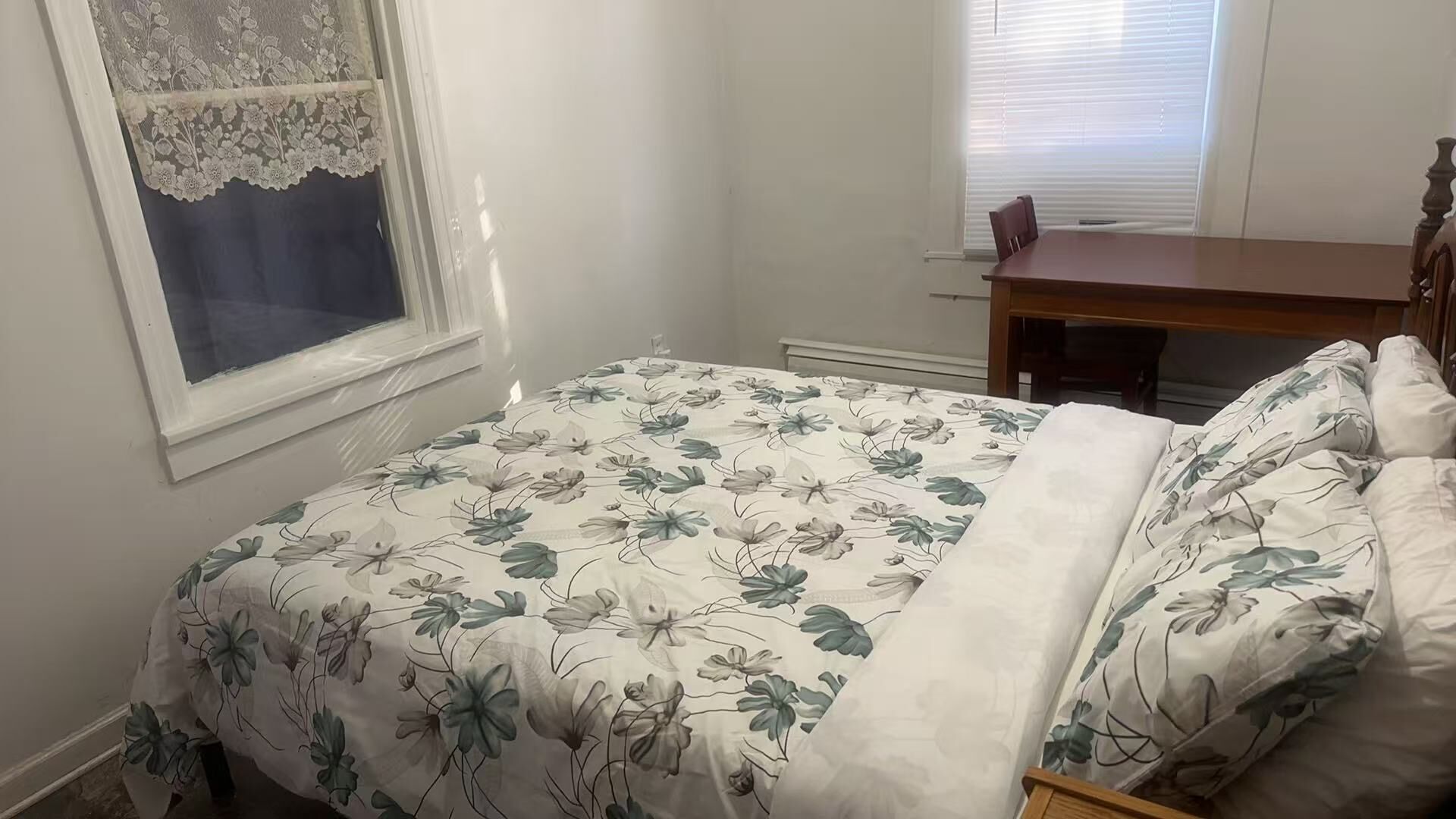 Une chambre à coucher avec une couvre-lit à motifs floraux, une table de chevet en bois, une chaise et une fenêtre ornée d’un rideau en dentelle.