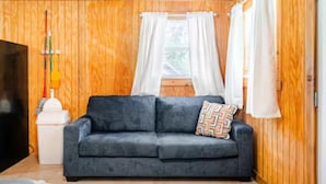 A room with a grey sofa, a small white table, and a window with white curtains. - Bixby Cove Cabin #3 (Porum)